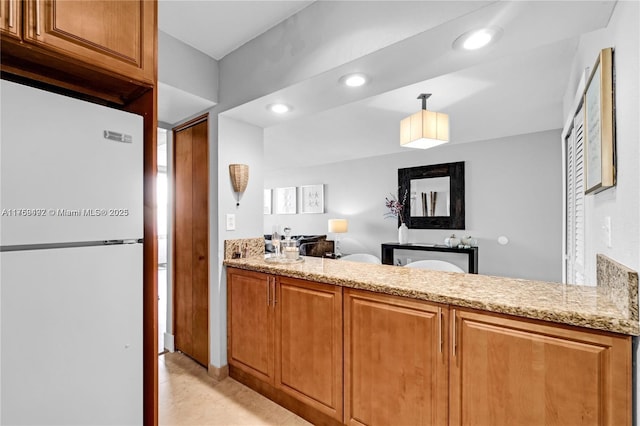 kitchen with recessed lighting, a peninsula, brown cabinets, freestanding refrigerator, and light stone countertops