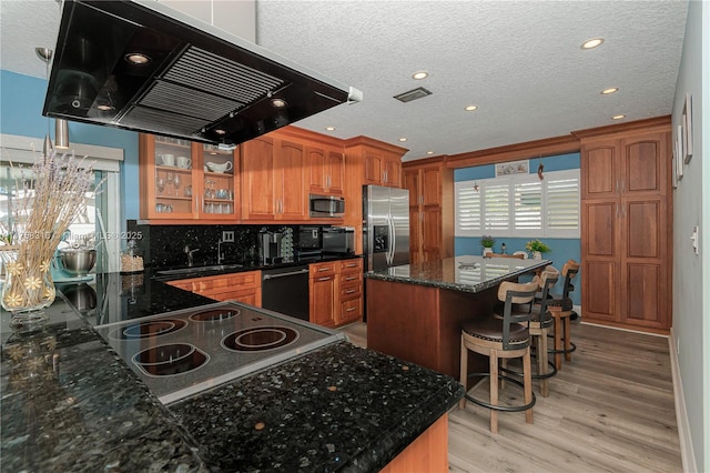 kitchen with range hood, appliances with stainless steel finishes, a center island, brown cabinetry, and glass insert cabinets