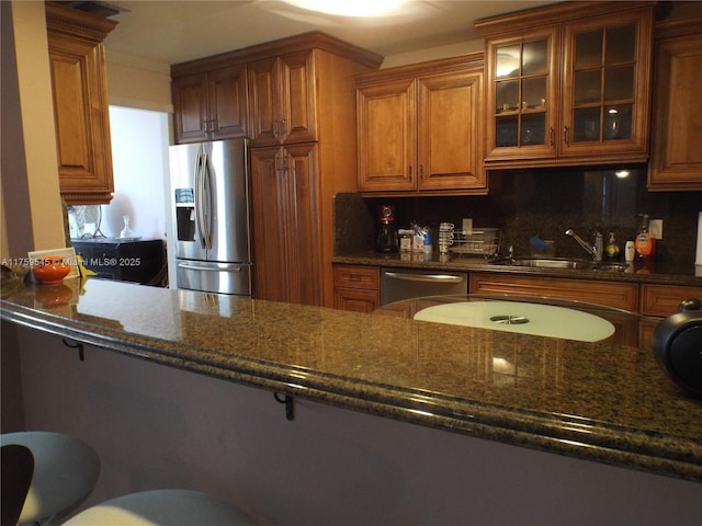 kitchen featuring stainless steel appliances, brown cabinetry, a sink, dark stone counters, and glass insert cabinets
