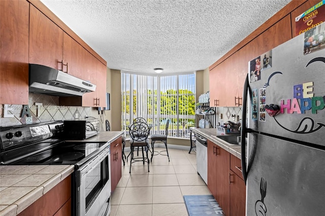 kitchen featuring brown cabinets, tile countertops, light tile patterned floors, appliances with stainless steel finishes, and under cabinet range hood