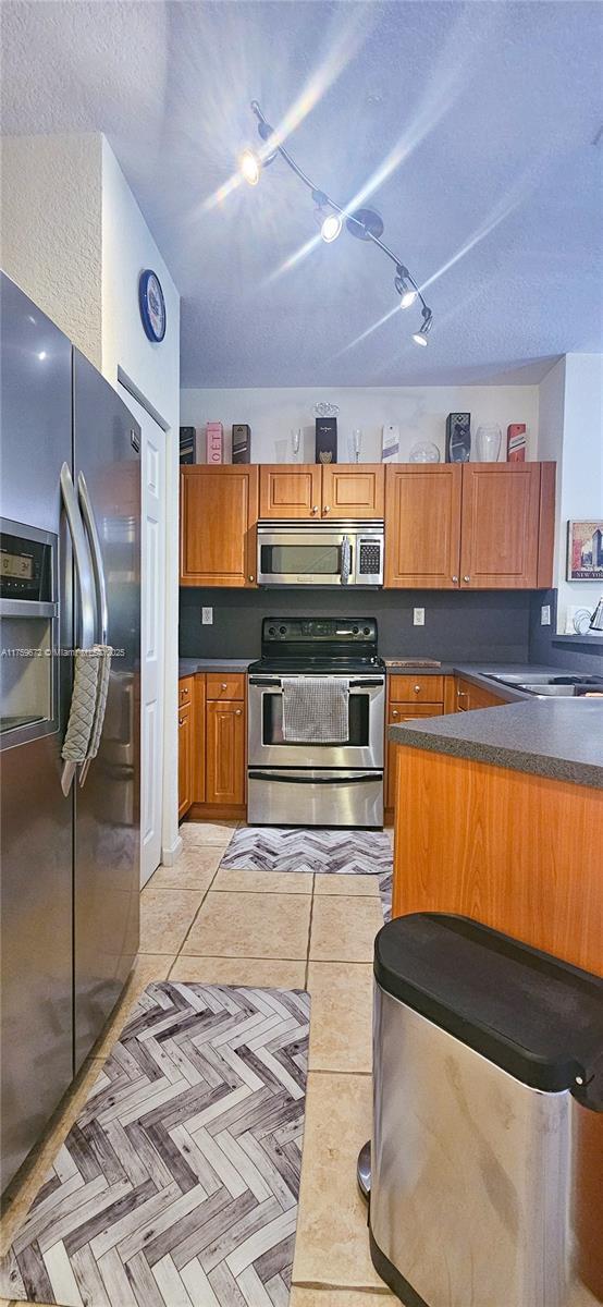 kitchen with light tile patterned flooring, a sink, appliances with stainless steel finishes, brown cabinetry, and dark countertops