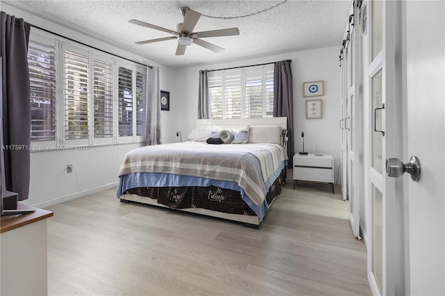 bedroom with a textured ceiling, ceiling fan, a barn door, light wood-style flooring, and baseboards