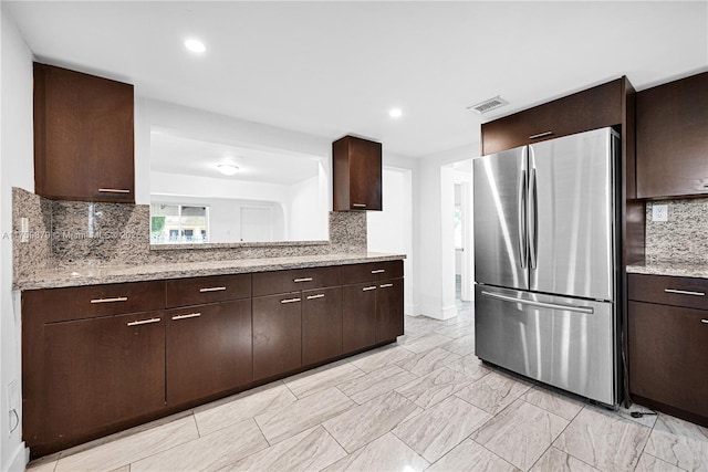 kitchen featuring light stone counters, visible vents, freestanding refrigerator, dark brown cabinets, and backsplash