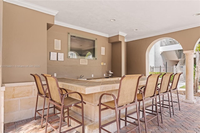 bar with indoor wet bar, crown molding, brick floor, and decorative columns