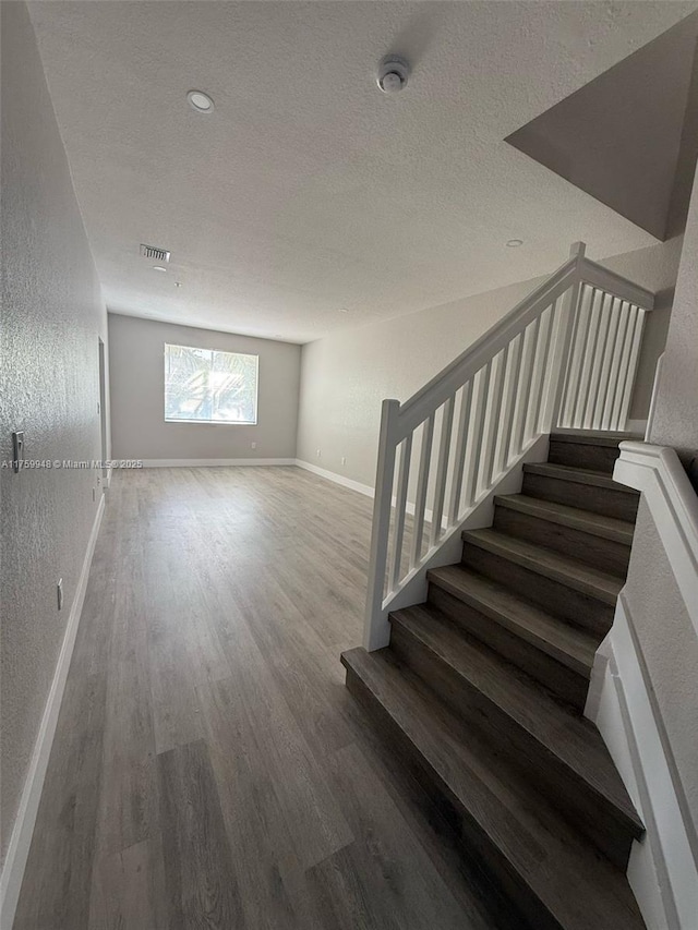 staircase featuring wood finished floors, visible vents, baseboards, a textured ceiling, and a textured wall