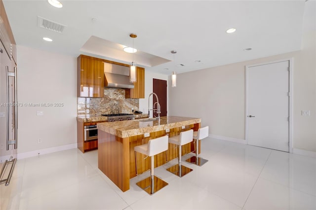 kitchen featuring visible vents, decorative backsplash, brown cabinets, wall chimney exhaust hood, and a raised ceiling