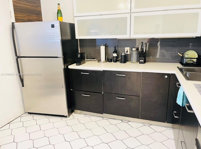 kitchen featuring freestanding refrigerator, a sink, light countertops, glass insert cabinets, and backsplash