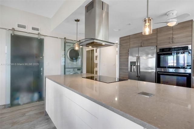 kitchen featuring double wall oven, island exhaust hood, stainless steel refrigerator with ice dispenser, stacked washer / dryer, and black electric cooktop