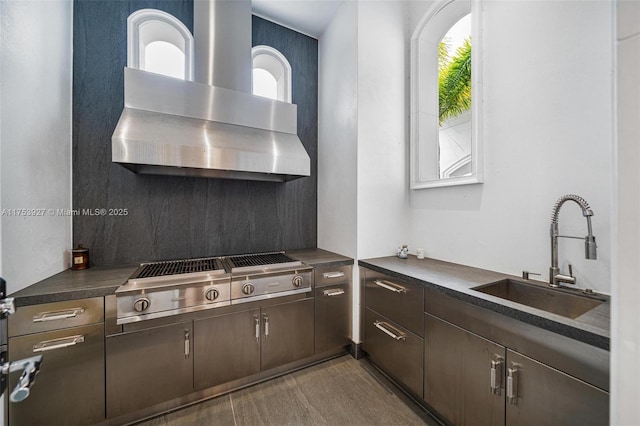 kitchen featuring a sink, dark countertops, wall chimney range hood, dark brown cabinets, and stainless steel gas cooktop