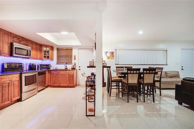 kitchen featuring brown cabinetry, a tray ceiling, stainless steel appliances, glass insert cabinets, and dark countertops