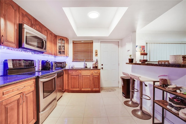 kitchen with light tile patterned floors, stainless steel appliances, glass insert cabinets, a raised ceiling, and brown cabinets