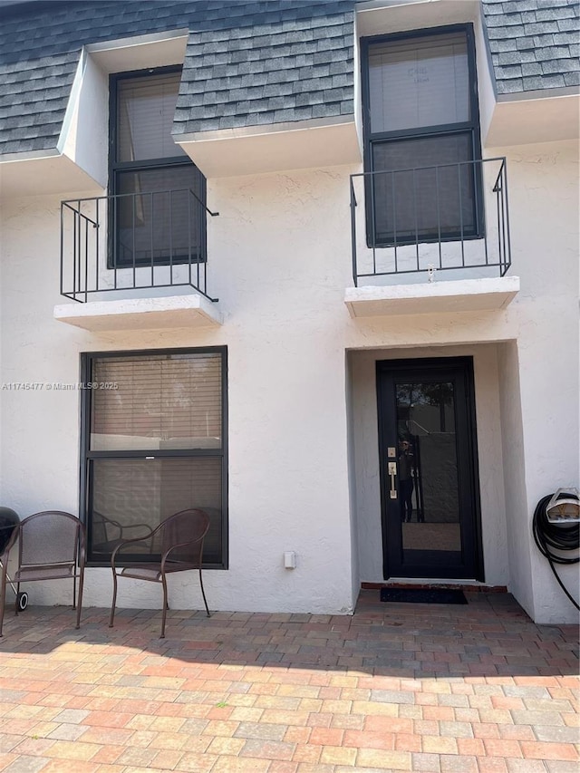 view of exterior entry featuring a balcony, stucco siding, a patio, and roof with shingles