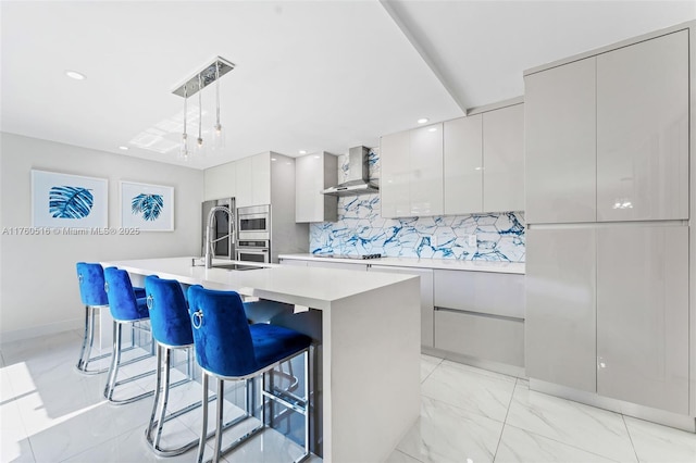 kitchen featuring wall chimney range hood, a center island with sink, light countertops, marble finish floor, and modern cabinets