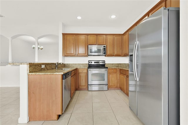 kitchen with brown cabinets, a sink, appliances with stainless steel finishes, light tile patterned flooring, and light stone countertops