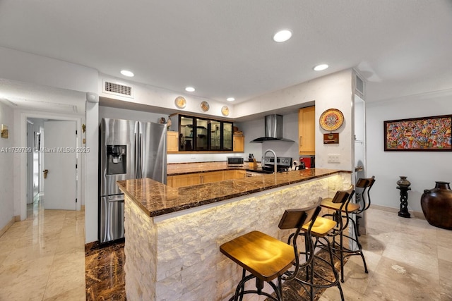 kitchen with a sink, dark stone counters, appliances with stainless steel finishes, a peninsula, and wall chimney range hood