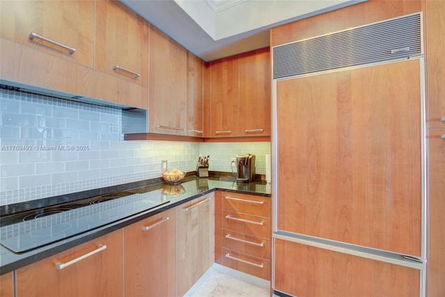 kitchen with dark stone countertops, brown cabinetry, built in refrigerator, black electric cooktop, and backsplash