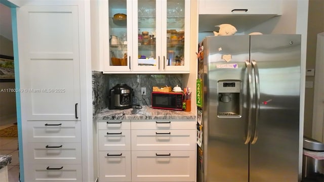 kitchen featuring light stone countertops, glass insert cabinets, stainless steel refrigerator with ice dispenser, white cabinetry, and backsplash