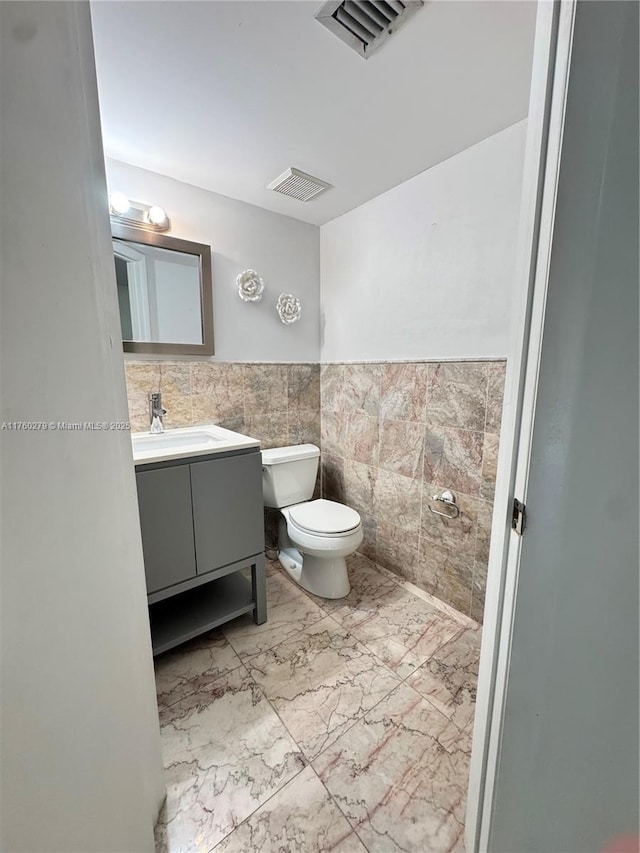 half bathroom featuring visible vents, tile walls, toilet, marble finish floor, and vanity