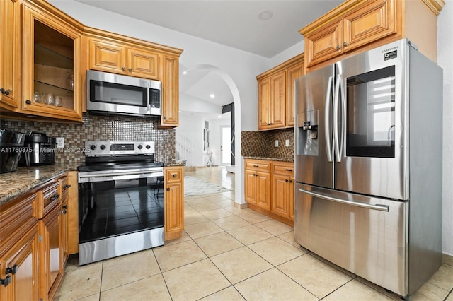 kitchen featuring dark stone countertops, light tile patterned flooring, stainless steel appliances, glass insert cabinets, and tasteful backsplash