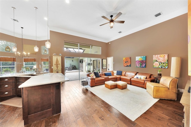 living area with visible vents, dark wood-style flooring, ornamental molding, a towering ceiling, and ceiling fan with notable chandelier