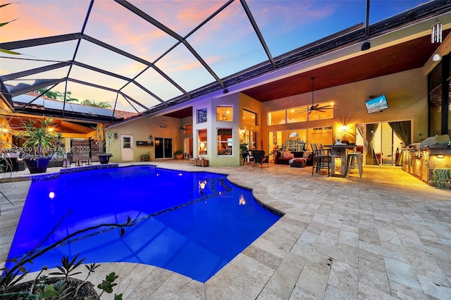 pool at dusk featuring an outdoor kitchen, glass enclosure, a patio, and ceiling fan