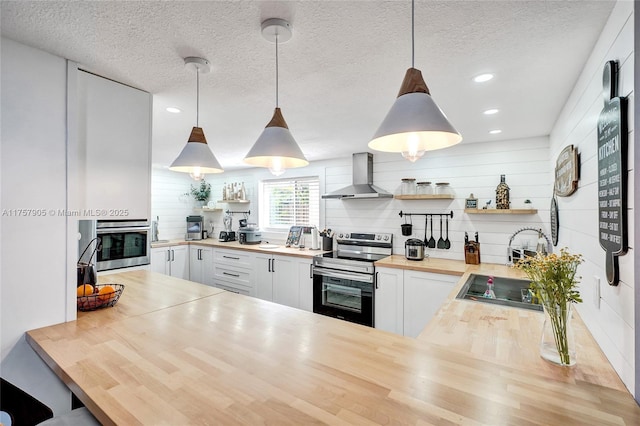 kitchen featuring open shelves, hanging light fixtures, appliances with stainless steel finishes, wall chimney exhaust hood, and butcher block counters