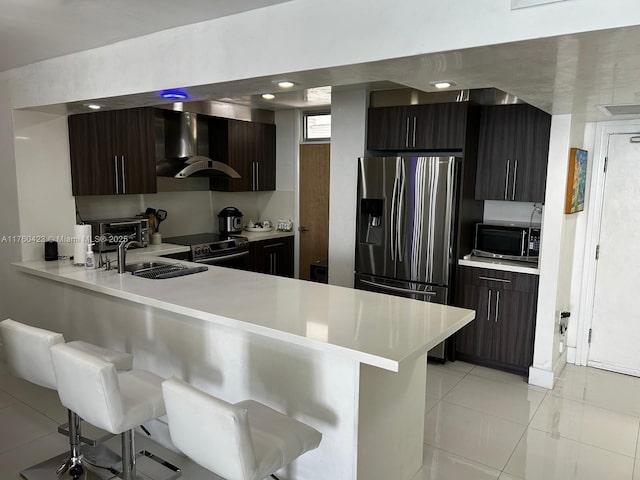 kitchen with a breakfast bar, stainless steel appliances, a sink, light countertops, and wall chimney exhaust hood