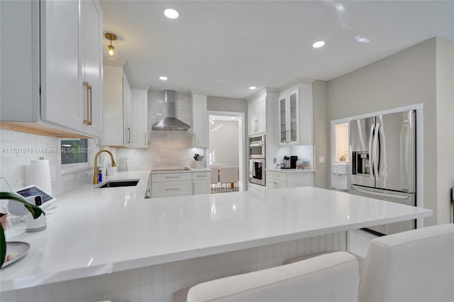 kitchen with a kitchen bar, a sink, stainless steel appliances, a peninsula, and wall chimney exhaust hood