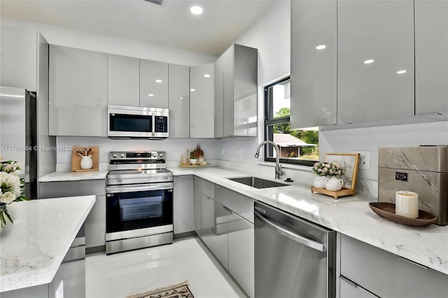 kitchen with modern cabinets, gray cabinetry, light stone counters, a sink, and appliances with stainless steel finishes