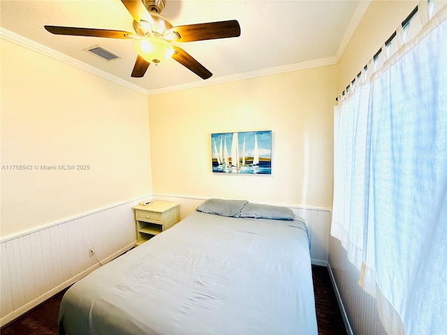 bedroom featuring a ceiling fan, crown molding, visible vents, and a wainscoted wall