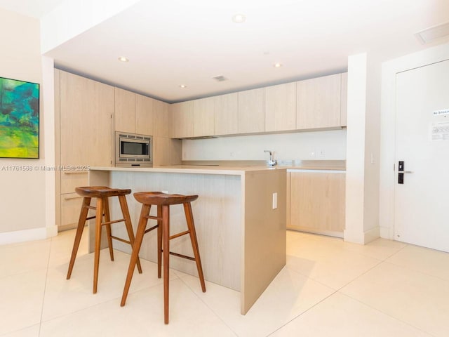 kitchen featuring a kitchen bar, stainless steel microwave, light countertops, and light brown cabinetry