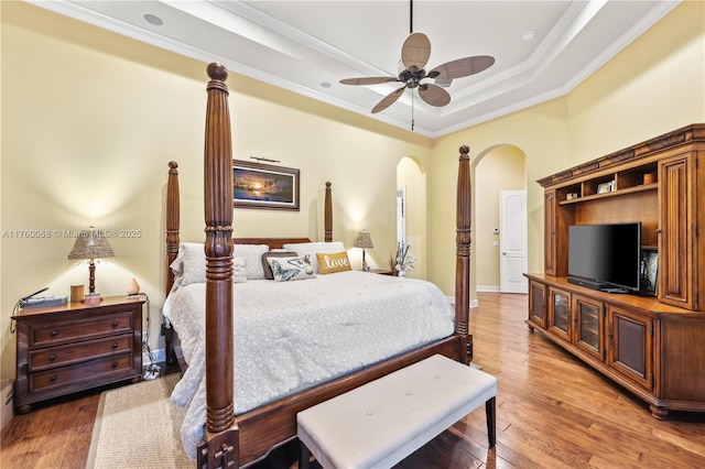 bedroom with a tray ceiling, crown molding, arched walkways, and light wood finished floors