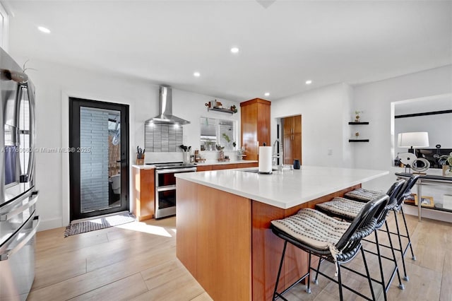 kitchen with light wood-style flooring, a sink, appliances with stainless steel finishes, brown cabinetry, and wall chimney range hood