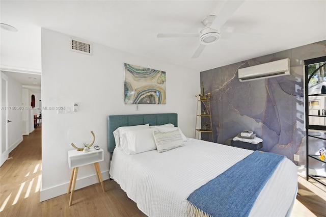 bedroom with a wall unit AC, a ceiling fan, wood finished floors, visible vents, and baseboards