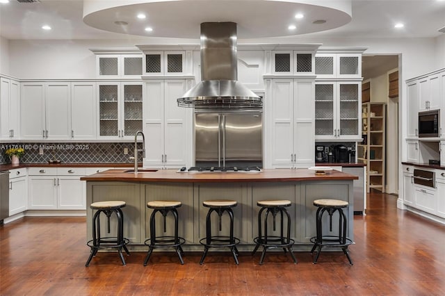 kitchen featuring built in appliances, island exhaust hood, dark wood-style floors, wood counters, and a kitchen island with sink