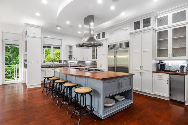 kitchen featuring a kitchen island with sink, appliances with stainless steel finishes, island exhaust hood, wood counters, and open shelves