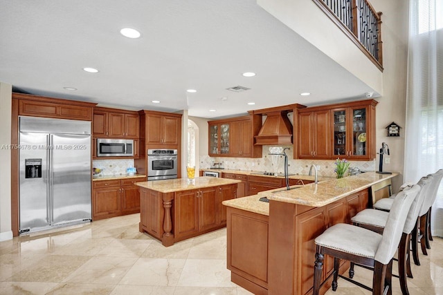 kitchen featuring premium range hood, visible vents, appliances with stainless steel finishes, brown cabinetry, and light stone countertops