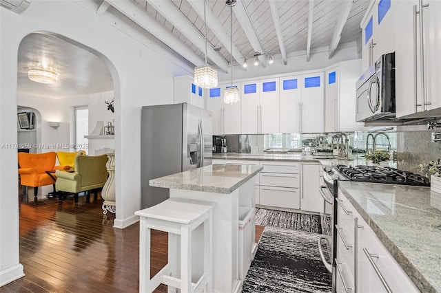 kitchen featuring beamed ceiling, light stone counters, backsplash, arched walkways, and appliances with stainless steel finishes