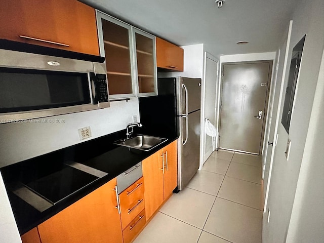 kitchen with a sink, dark countertops, appliances with stainless steel finishes, light tile patterned floors, and glass insert cabinets