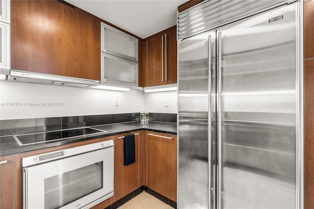 kitchen with glass insert cabinets, oven, black electric stovetop, under cabinet range hood, and built in refrigerator