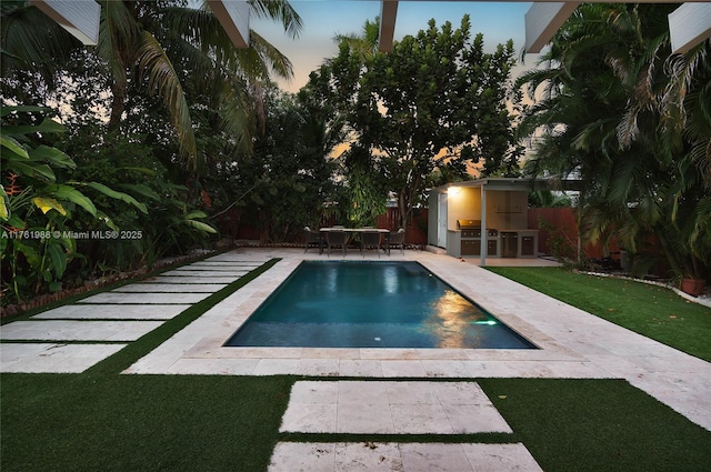 pool at dusk with outdoor dry bar, fence, a yard, area for grilling, and a patio