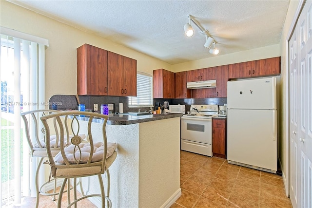 kitchen featuring under cabinet range hood, backsplash, dark countertops, white appliances, and a peninsula