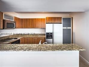 kitchen featuring baseboards, a peninsula, brown cabinetry, stone countertops, and stainless steel appliances