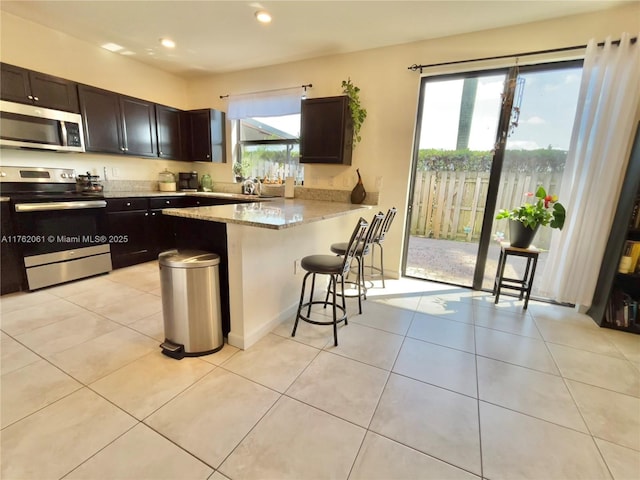 kitchen featuring dark brown cabinetry, a kitchen bar, light tile patterned floors, light stone counters, and appliances with stainless steel finishes