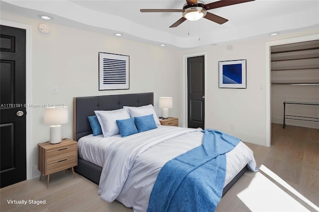 bedroom with light wood finished floors, a tray ceiling, recessed lighting, ceiling fan, and a walk in closet