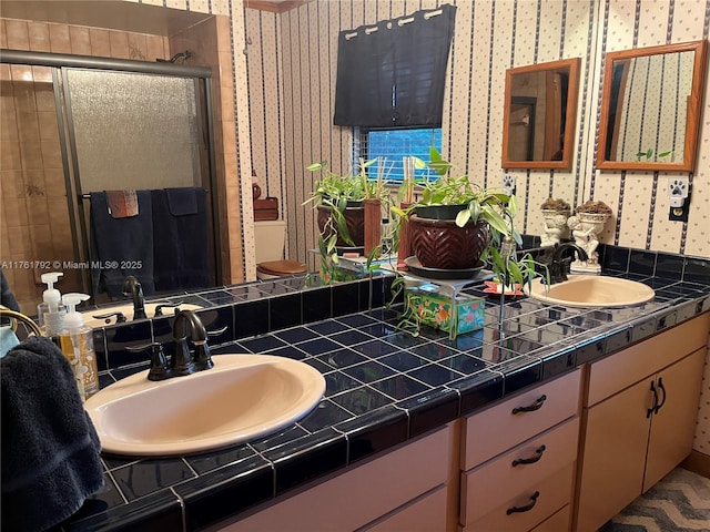 bathroom with wallpapered walls, a shower stall, double vanity, and a sink