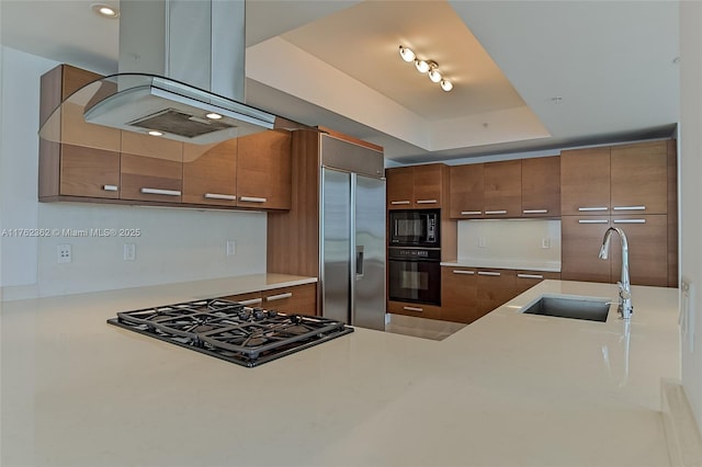 kitchen with black appliances, a sink, a tray ceiling, brown cabinetry, and island range hood