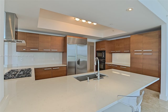 kitchen featuring island exhaust hood, brown cabinetry, black appliances, a raised ceiling, and a sink