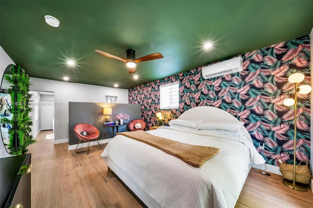 bedroom with wallpapered walls, baseboards, a wall unit AC, wood finished floors, and a ceiling fan