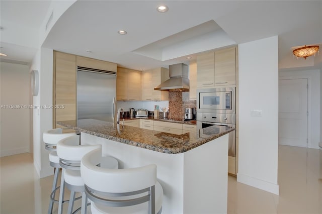 kitchen with a breakfast bar, dark stone counters, wall chimney range hood, decorative backsplash, and built in appliances